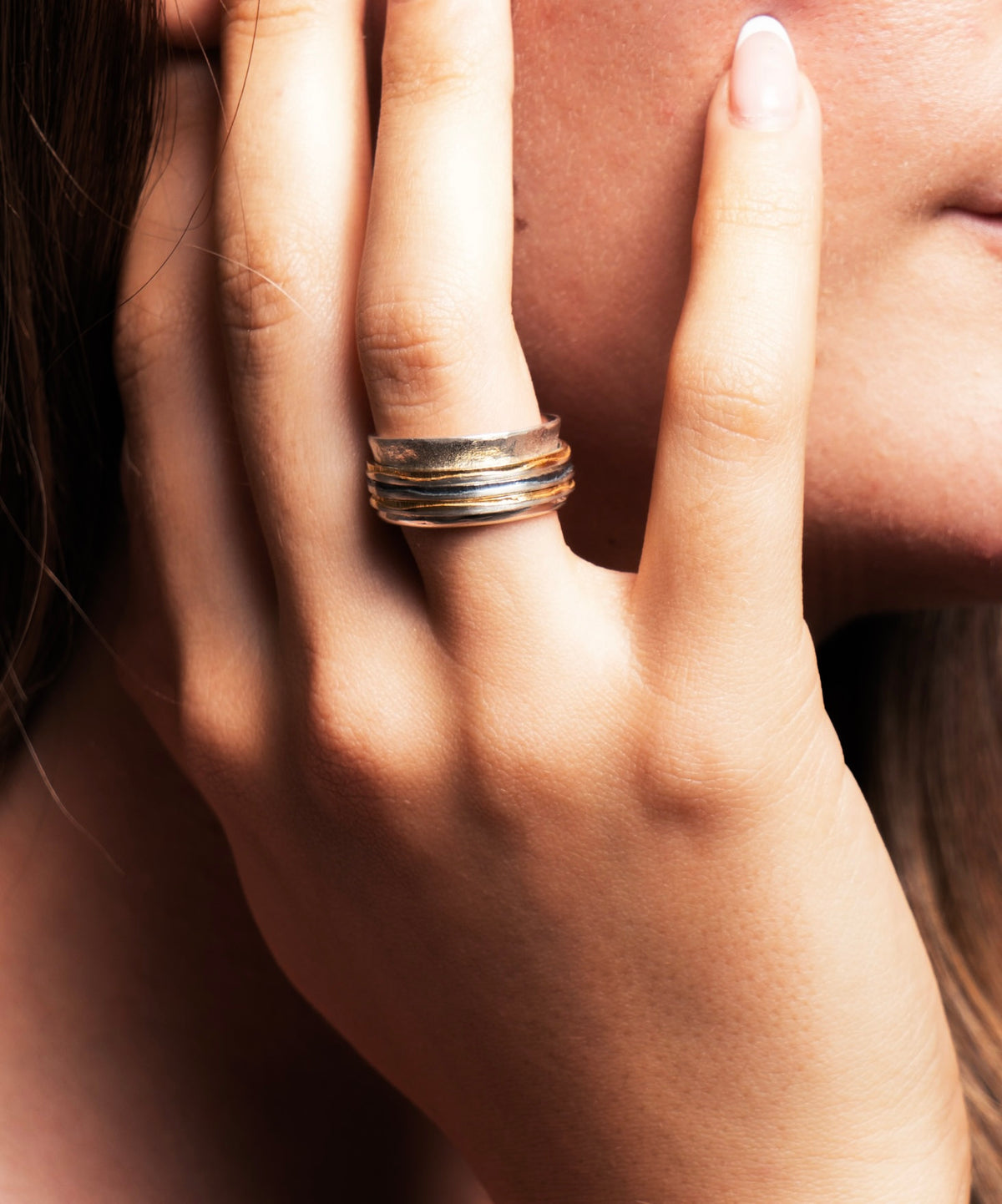 A close-up of a person's hand wearing a stack of three thin metallic rings, with the face partially visible in the background.
