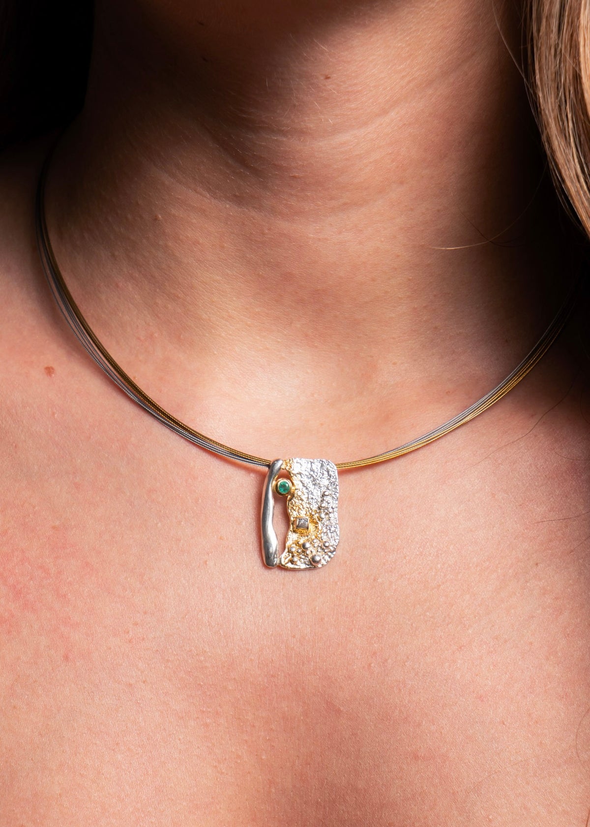 A close-up of a model's neck and chest wearing a delicate gold-tone cable necklace with a textured, irregular-shaped silver pendant featuring a small green stone and yellow accent.