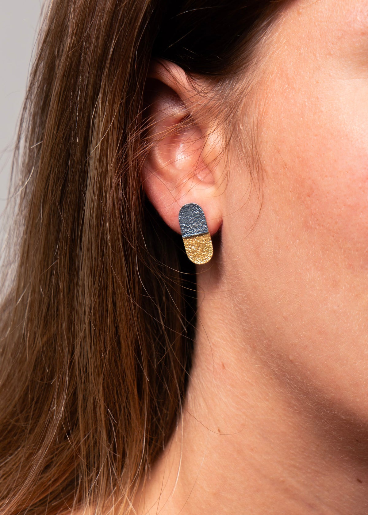A close-up of a model's ear wearing a small capsule-shaped stud earring with a textured dark blue top half and a gold bottom half.