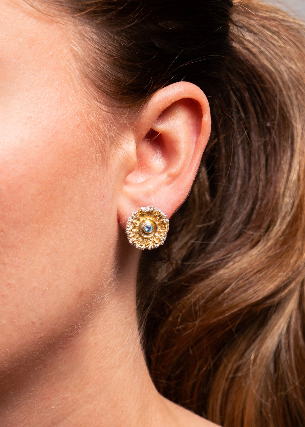 A close-up of a person's ear wearing a textured gold stud earring with a central round blue gemstone.