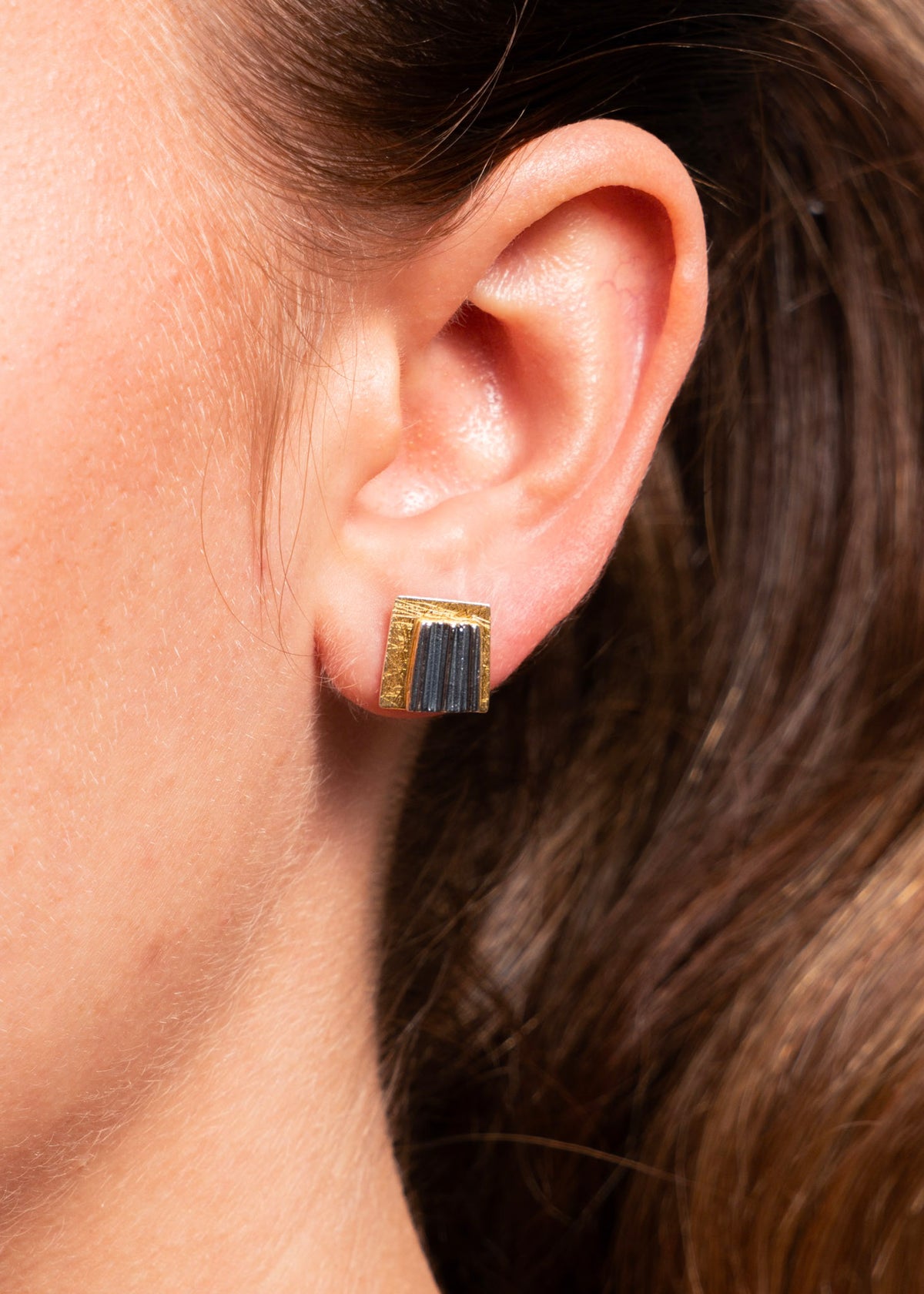 Close-up of a person's ear wearing a small square stud earring with vertical blue-gray inlay and gold-toned edges.