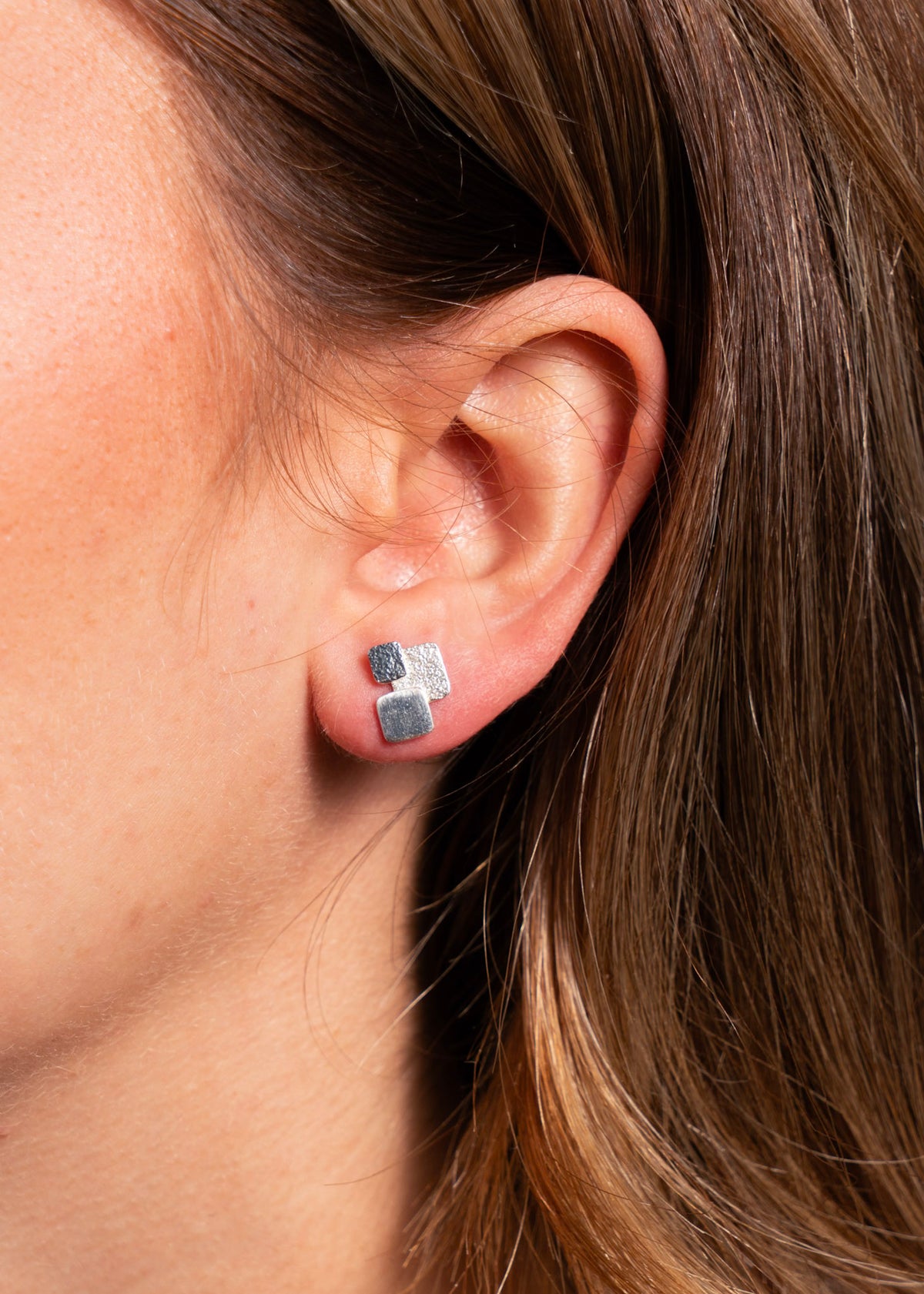 A close-up of a person's ear wearing a pair of silver stud earrings with textured square and rectangular charms.