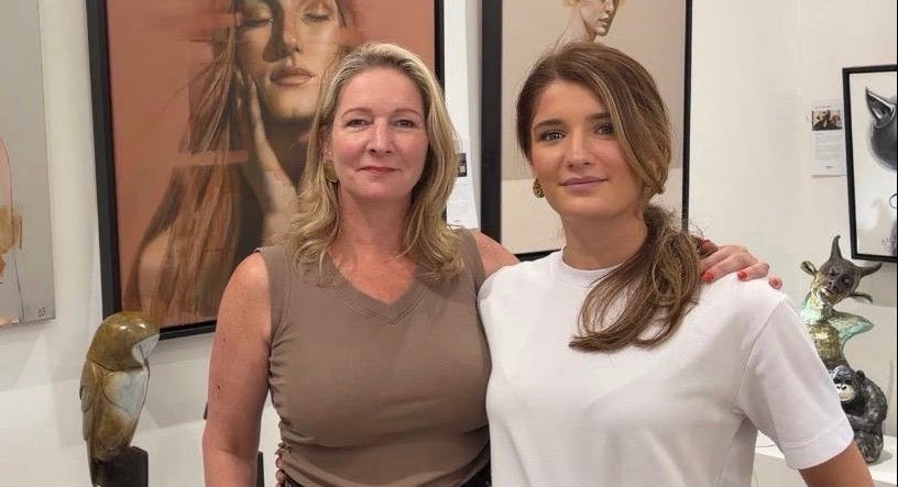 Two women standing together in an art gallery, with framed portraits of women hanging on the wall behind them and small sculptures displayed nearby.