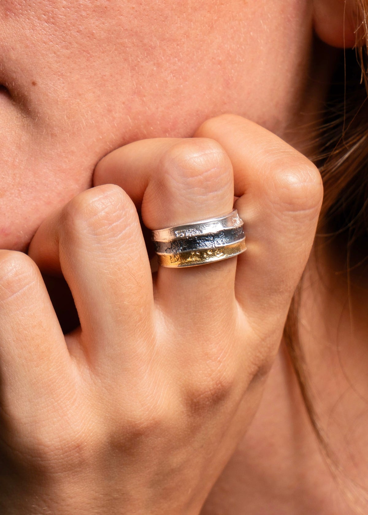 A close-up of a person's hand and cheek wearing a textured two-tone ring with silver and gold bands.