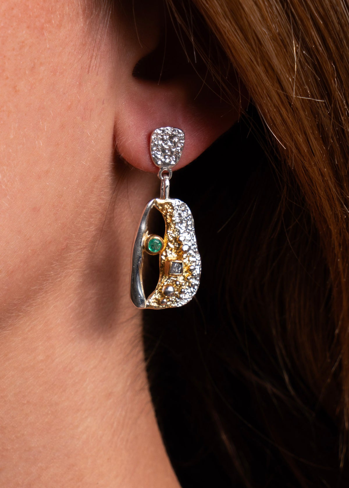 A close-up of a model's ear wearing a dangling earring. The earring features a silver-toned top setting with clear stones, a gold-toned curved drop with pavé-set clear stones, and a small green oval accent stone.