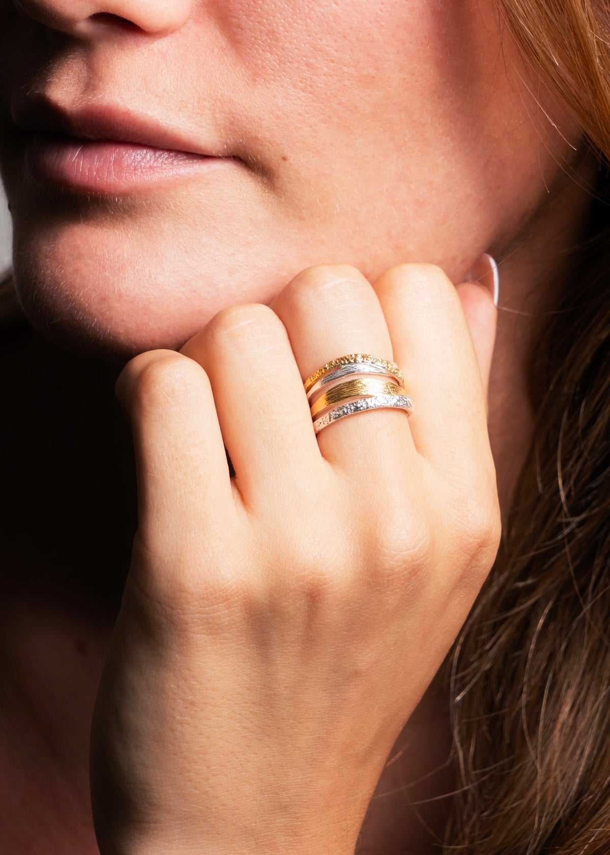 A close-up of a woman's hand resting near her chin, wearing two stacked rings: one with a gold band and the other with a silver band set with small clear stones.