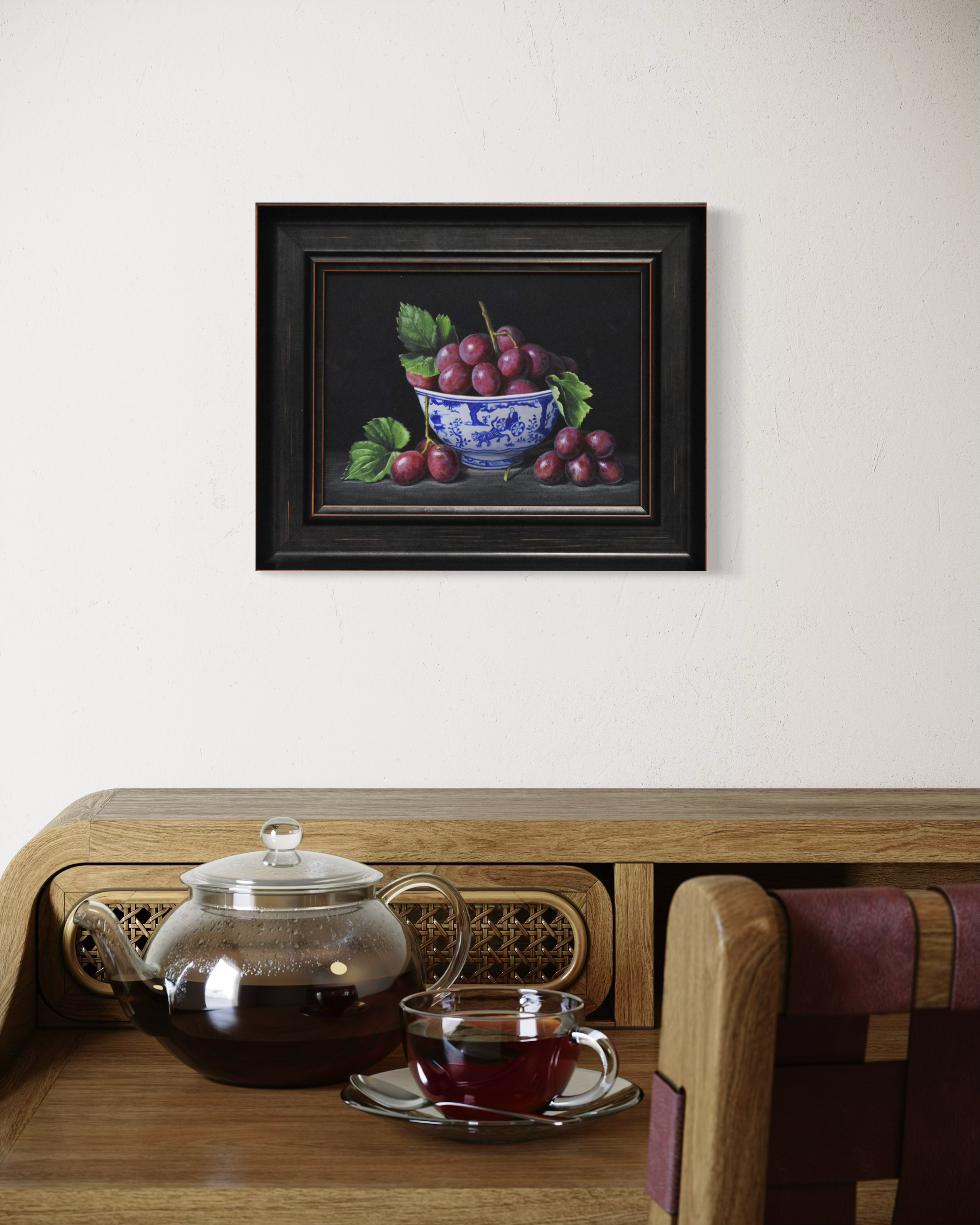 Framed still life painting of cherries on a wall above a wooden table with a teapot and cup.