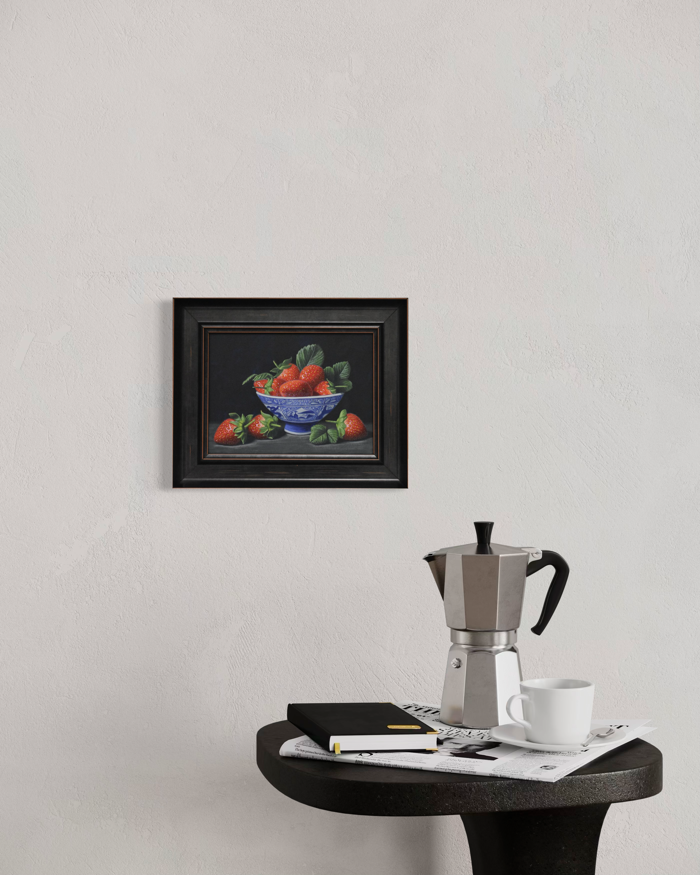 Framed still life painting on a wall above a small round table with a coffee maker and cup.