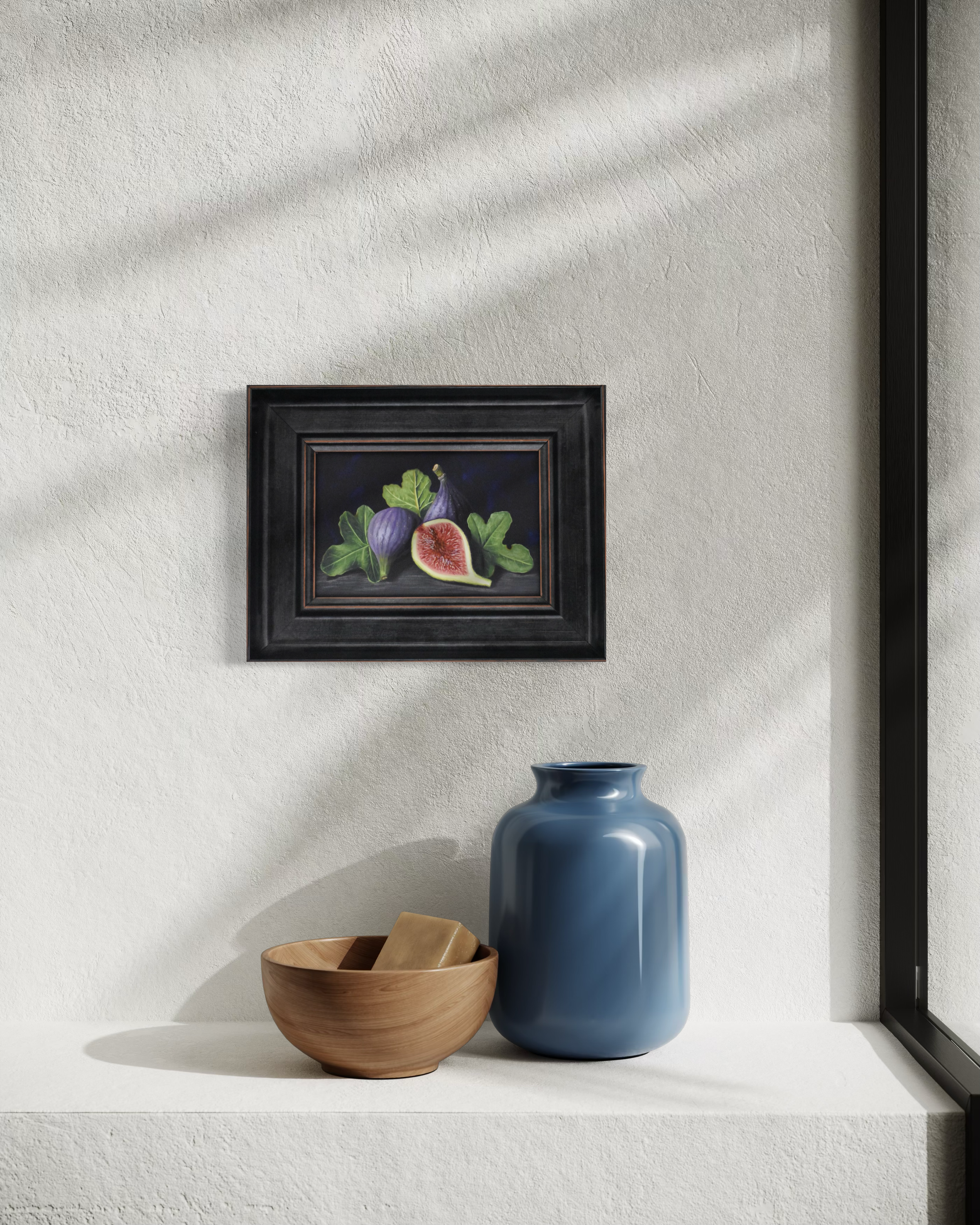 Framed artwork of figs on a wall with a blue vase and wooden bowl below.