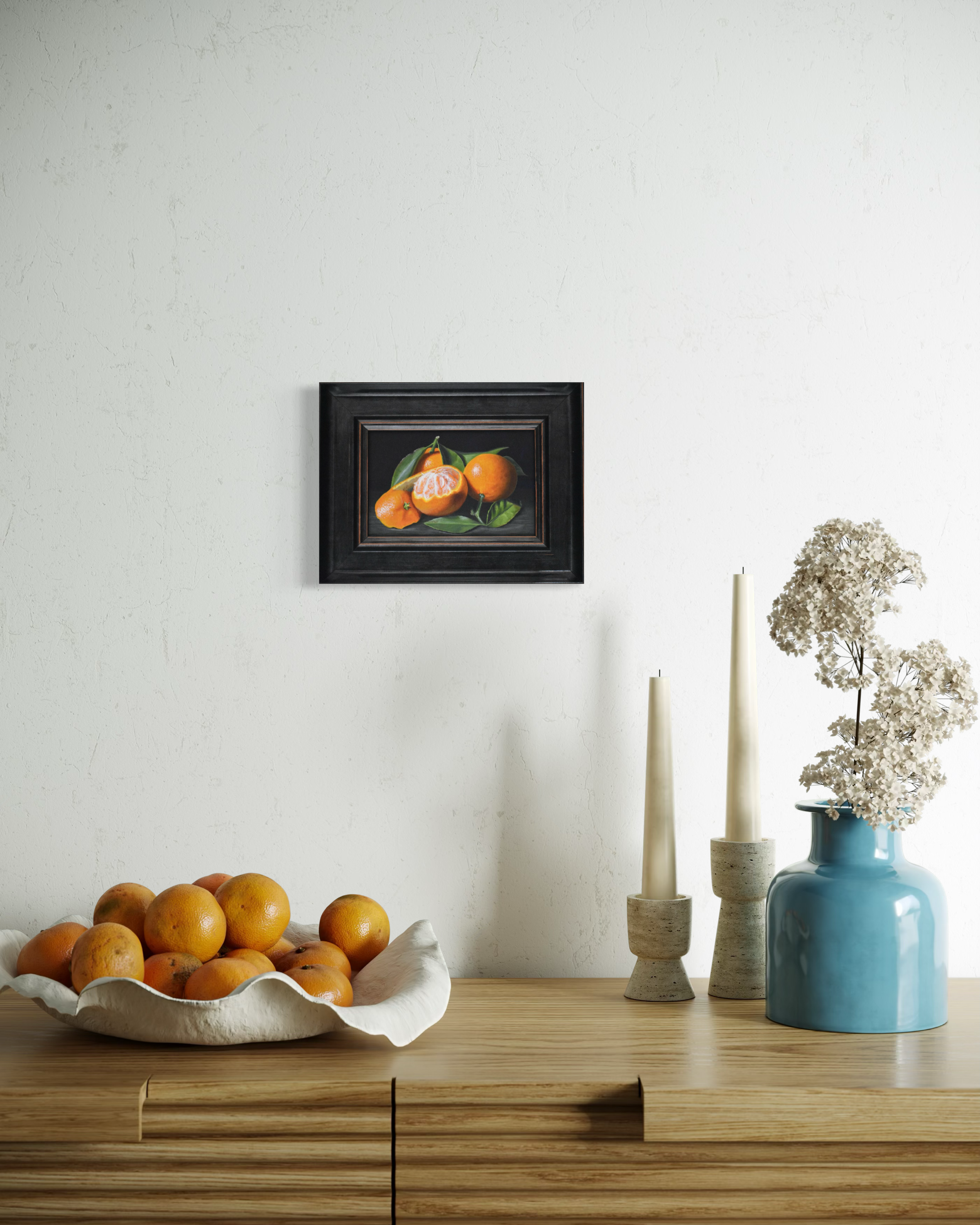 Decorative setup with a bowl of oranges, a vase with flowers, and a framed artwork on a wall.